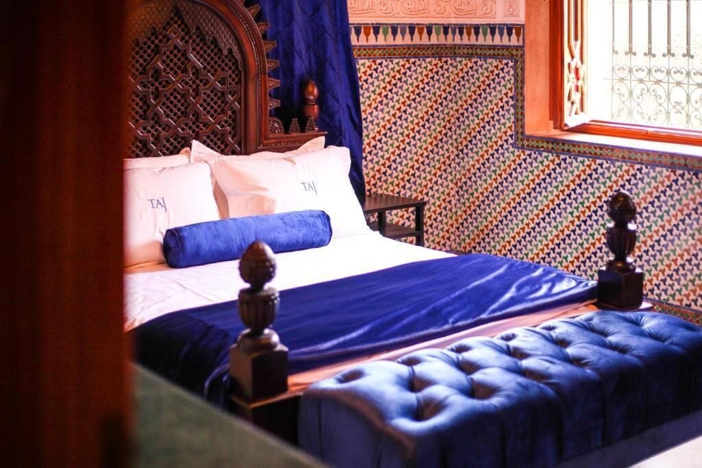 A bed with a blue headboard and pillows, a wooden headboard with intricate carvings, a patterned wall, and a window with metal bars.