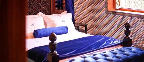 Una cama con cabecera azul y almohadillas, cabecera de madera con detalles tallados, pared estampada y ventana con barrotes.