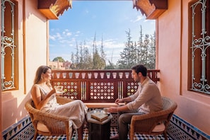 A balcony with wicker furniture, a small table with a book and a cup, and a view of trees and buildings. - Villa Taj Sofia & Spa (Marrakech)