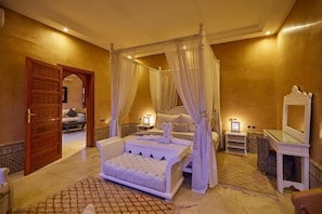 Ein luxuriöses Schlafzimmer mit einem Himmelbett, einer Sitzecke mit einer gepolsterten Couch, einem Schminktisch mit Spiegel und einer traditionellen Holztür.