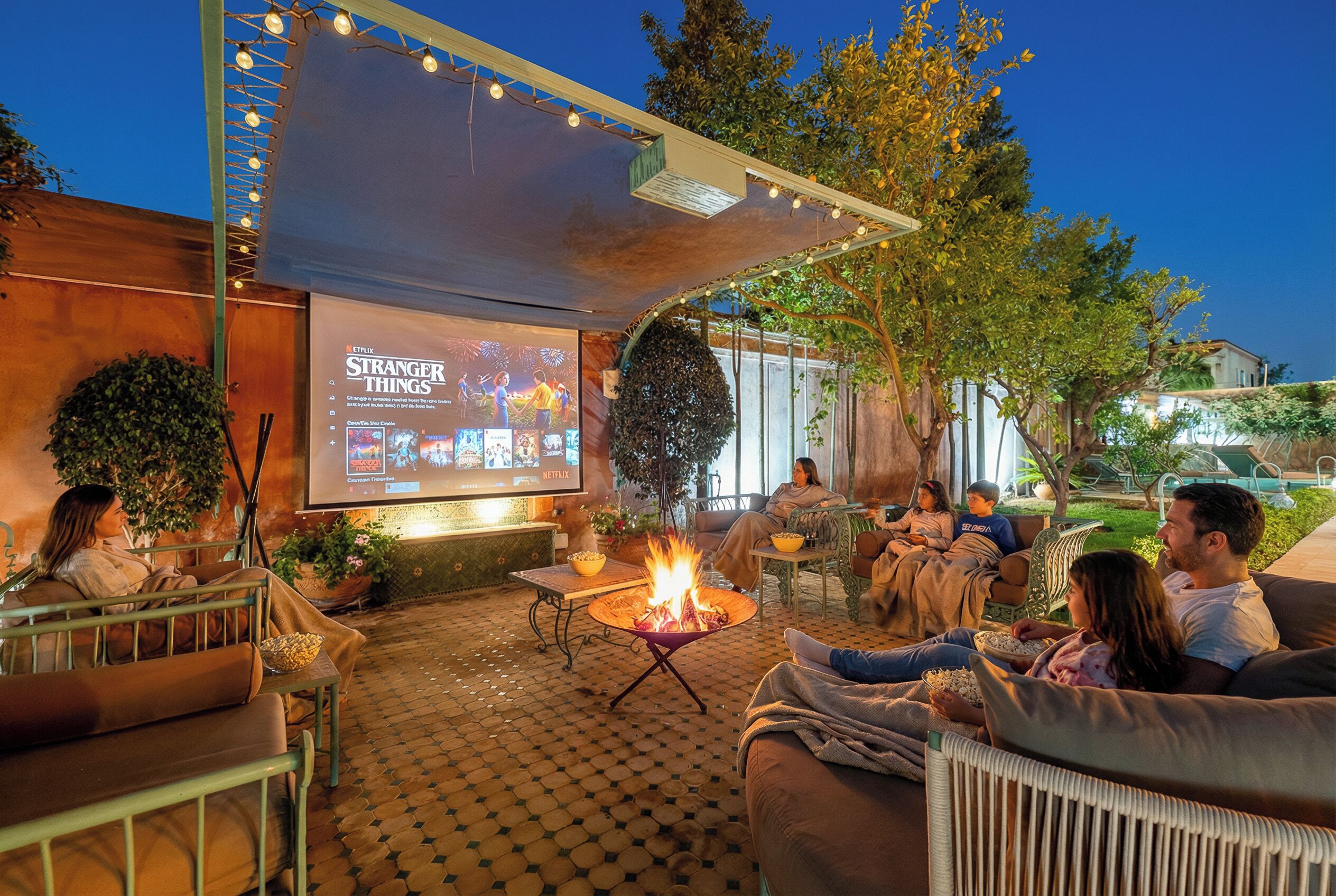 An outdoor seating area with a fire pit, a projection screen displaying a show, and a relaxed atmosphere.