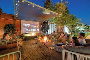 An outdoor seating area with a fire pit, a projection screen displaying a show, and a relaxed atmosphere.