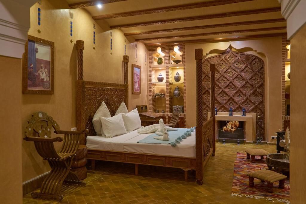 Quarto tradicional com cama, uma cadeira de madeira, lareira e prateleiras decorativas na parede.