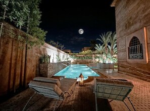 A pool area with lounge chairs and a lantern, surrounded by a brick wall and palm trees, under a night sky with a visible moon. - Villa Taj Sofia & Spa (Marrakech)