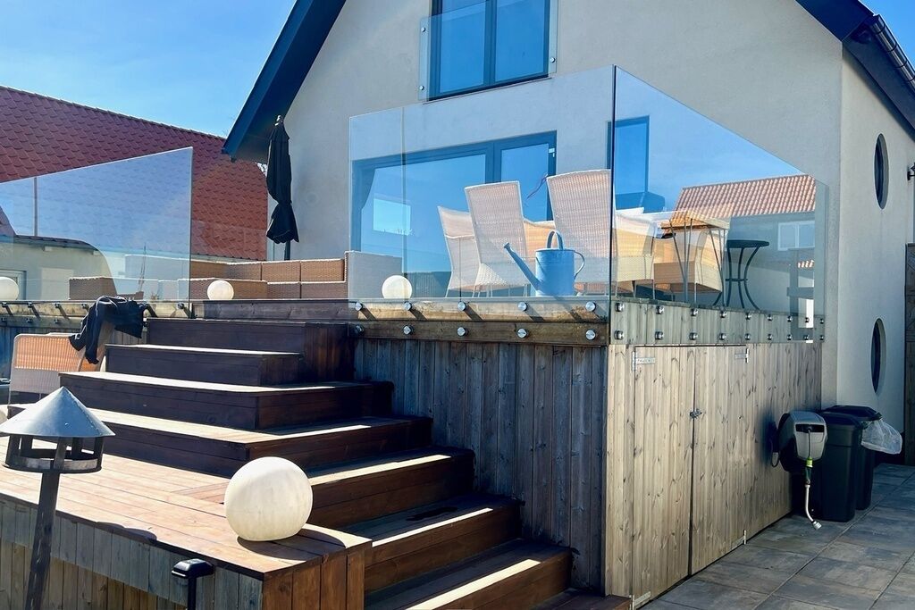Eine moderne Außenterrasse mit Holzstufen, die zu einem Glasgeländer führen, einem Sitzbereich mit Stühlen und einem Tisch sowie einem Mülleimer.