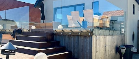 A modern outdoor deck with wooden steps leading to a glass balustrade, a seating area with chairs and a table, and a trash bin.