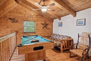 Un interior de madera con mesa de billar, decoración con estrellas, ventilador de techo y ventana con persianas.
