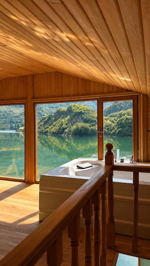Interior de una cabaña de madera con grandes ventanales que dan a un lago y un bosque.