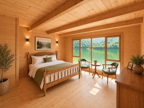 Un dormitorio tipo cabaña de madera con una cama, sillas, una mesita y una ventana con vista a un lago.