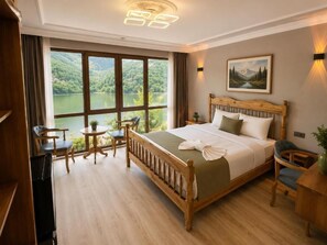 A hotel room with a large bed, a desk, a chair, a small table, and a view of a lake through large windows.