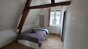 Une chambre avec un plafond en pente, un lit avec une couverture, un radiateur et une fenêtre avec des rideaux.