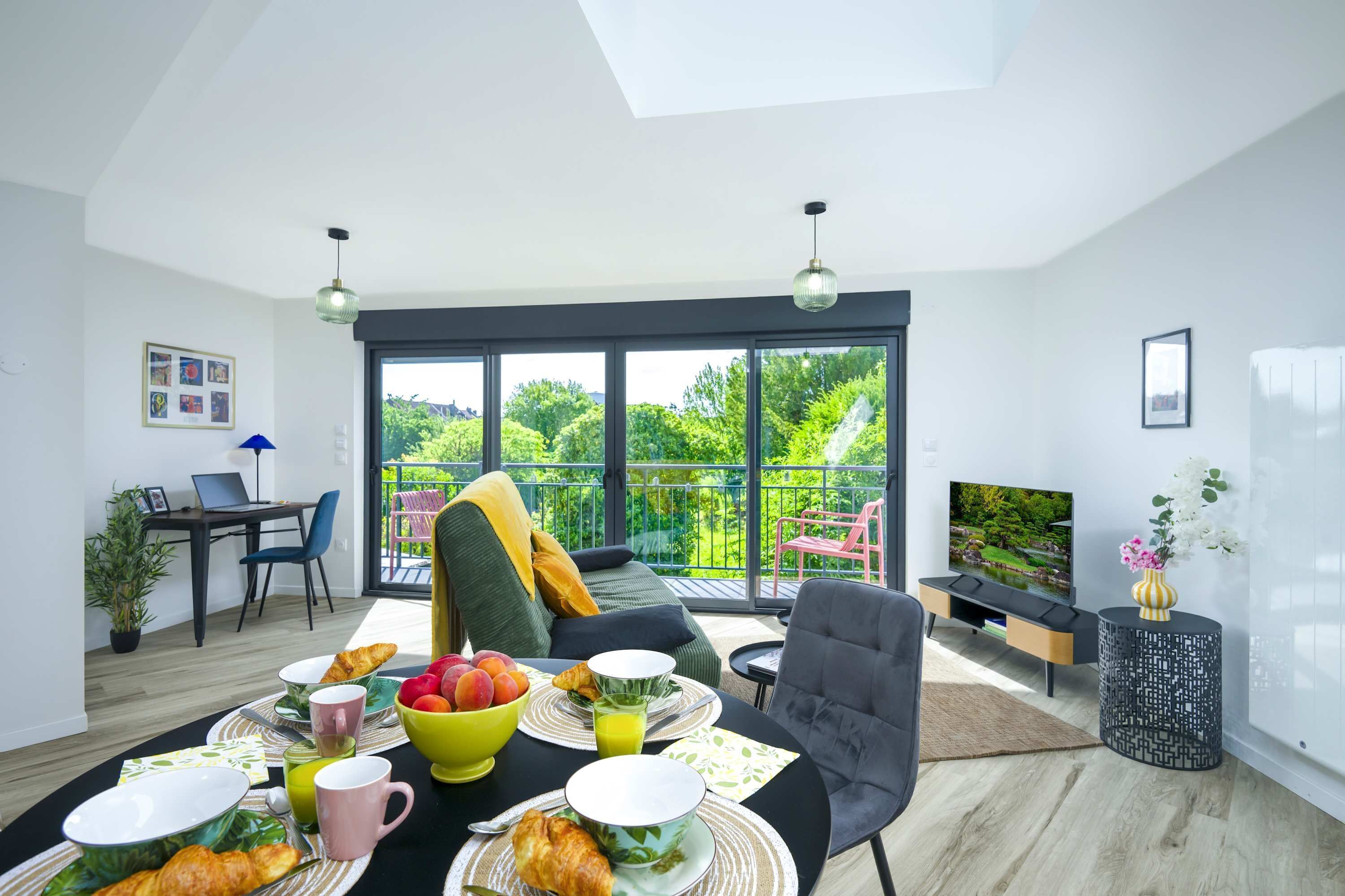 A modern living room with a dining area, a sofa, a TV, and a balcony with a view of greenery.