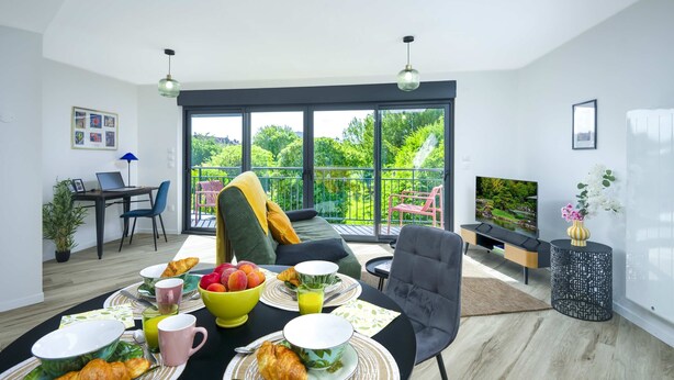 Ein modernes Wohnzimmer mit Essbereich, einer Couch, einem Fernseher und einem Balkon mit Blick auf Grünflächen.