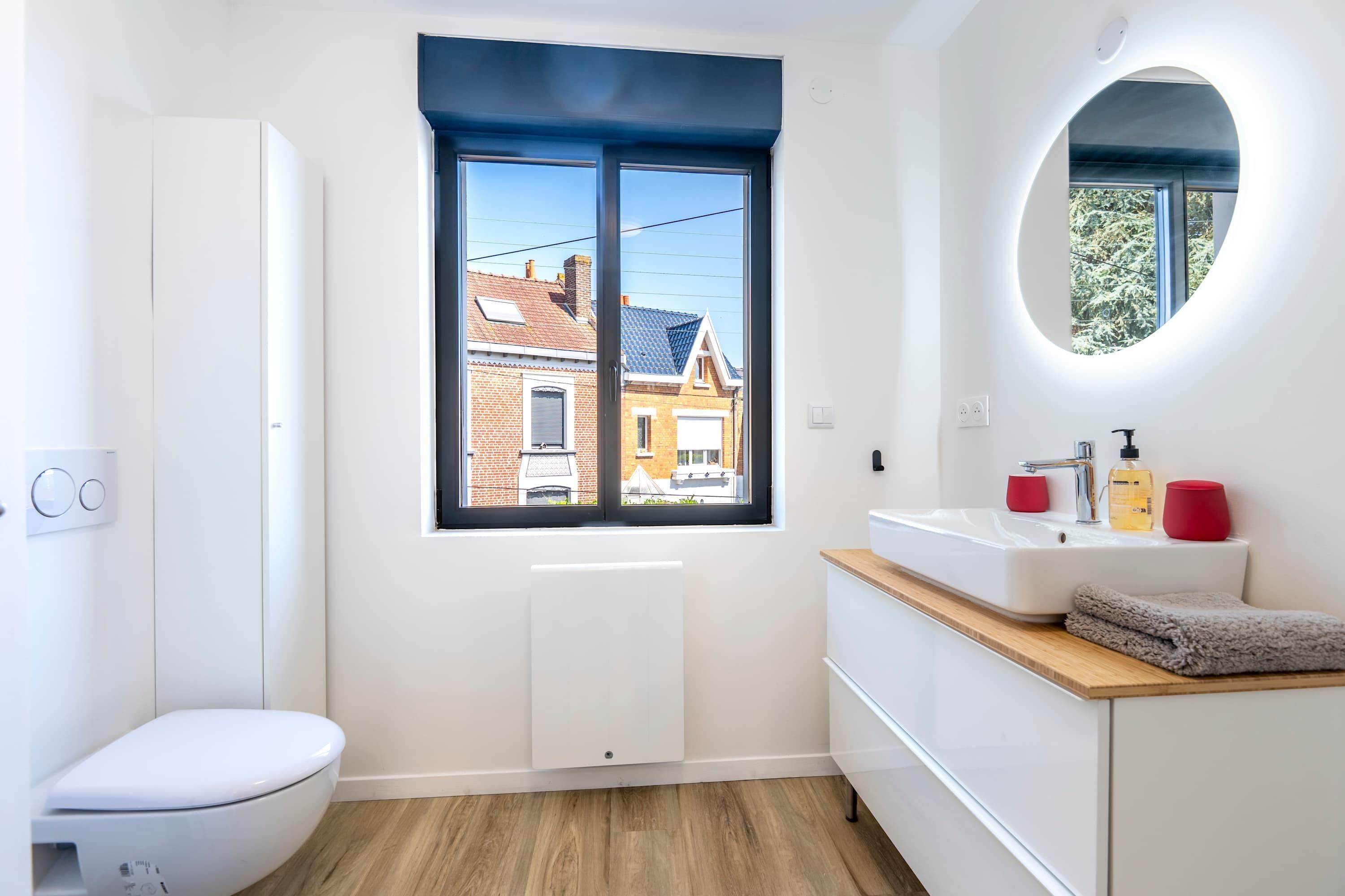 Un baño moderno con inodoro, un lavamanos con espejo y una ventana con vistas a una casa.