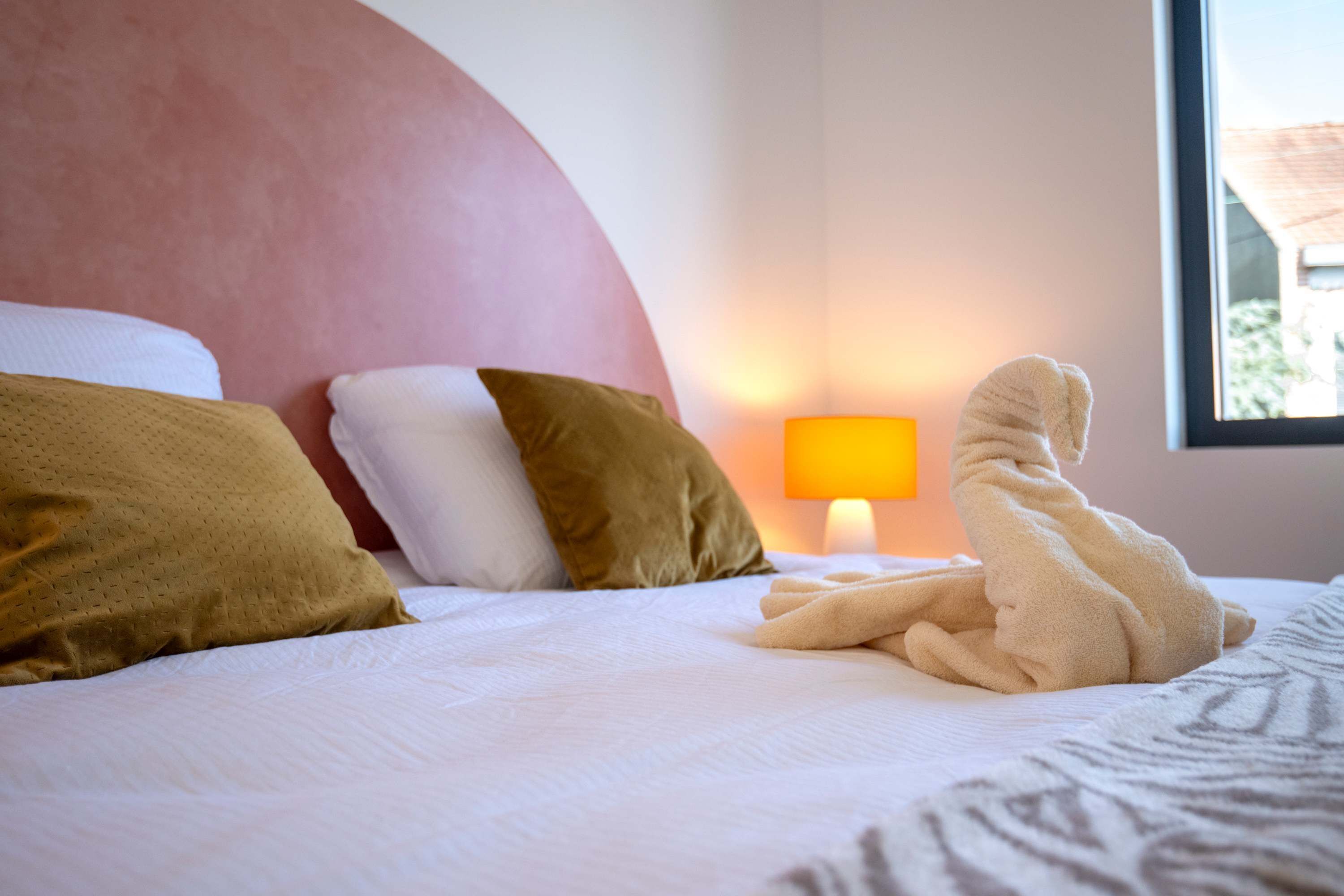 A hotel room with a bed, pillows, a lamp, and a window.