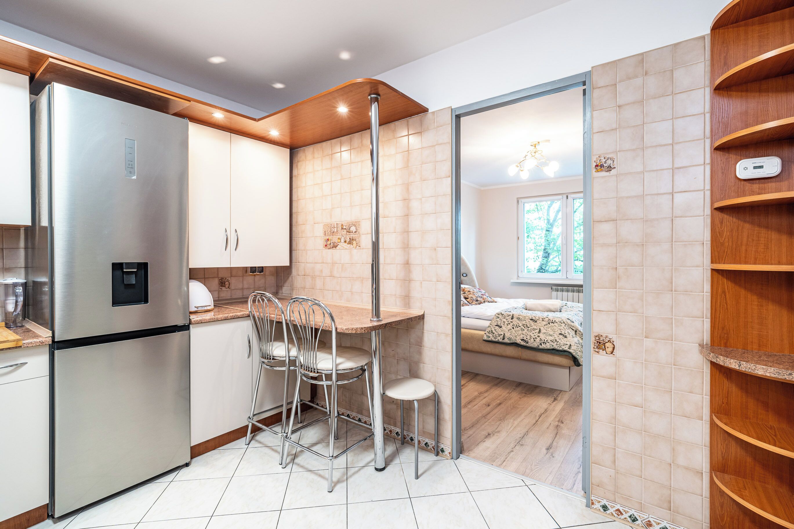 A modern kitchen with a built-in refrigerator, a dining area with metal chairs, and a view into a bedroom through a glass partition.