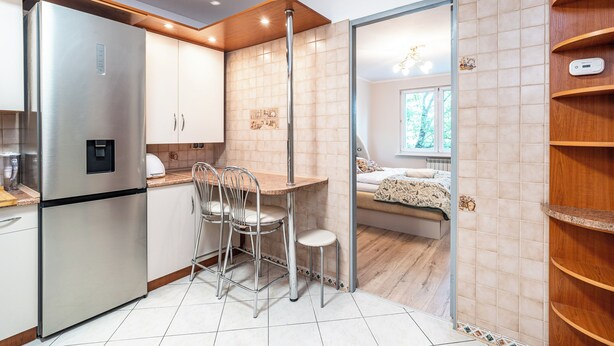 A modern kitchen with a built-in refrigerator, a dining area with metal chairs, and a view into a bedroom through a glass partition. - RentPlanet - Apartament Weissa (Kraków)