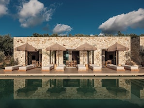 A poolside area with lounge chairs and umbrellas, stone walls, and a clear sky.