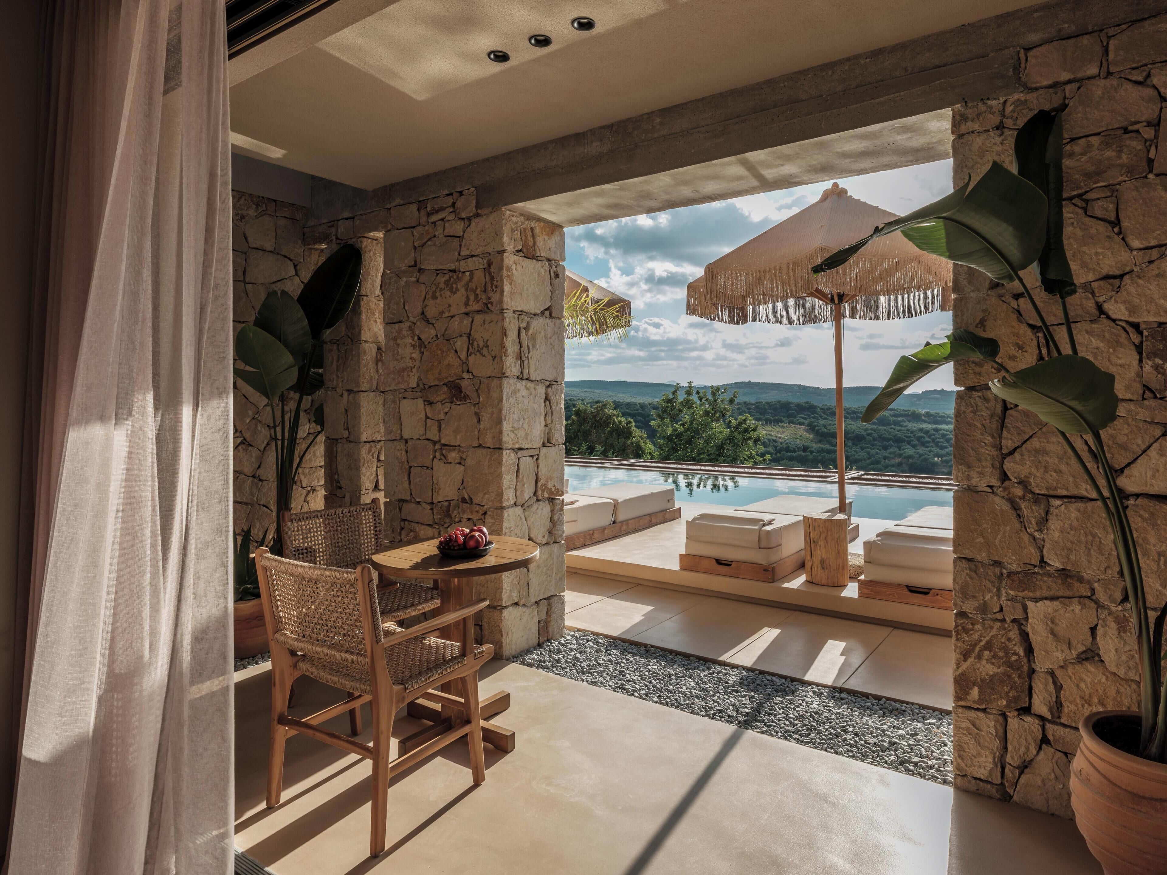 A room with a stone wall, a table with a bowl of fruit, a wicker chair, and a view of a pool and landscape.
