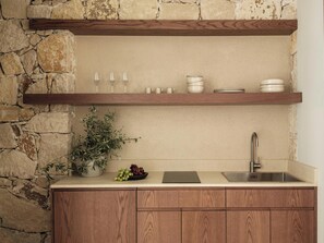 A kitchen with a stone wall, wooden cabinets, a countertop, a sink, and a shelf with glassware and plates. - Filema Suites (Xirokampi)