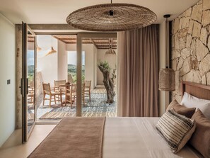 A bedroom with a bed, a dining area with chairs, a view through a glass door, and a stone wall.