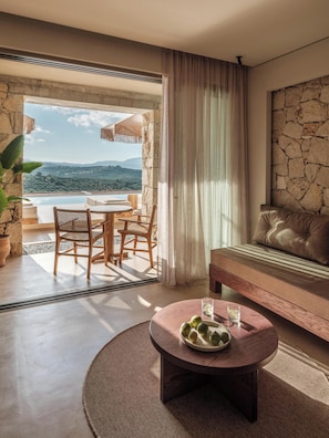 A room with a stone wall, a sofa, a round table with a plate of fruit, and a view of a pool and mountains. - Filema Suites (Xirokampi)