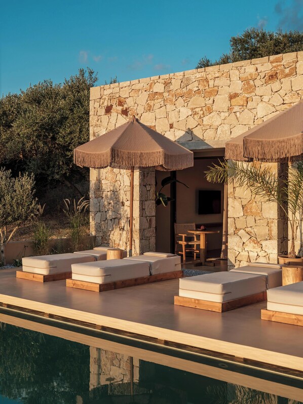 A poolside area with stone walls, lounge chairs, and umbrellas. - Filema Suites (Xirokampi)