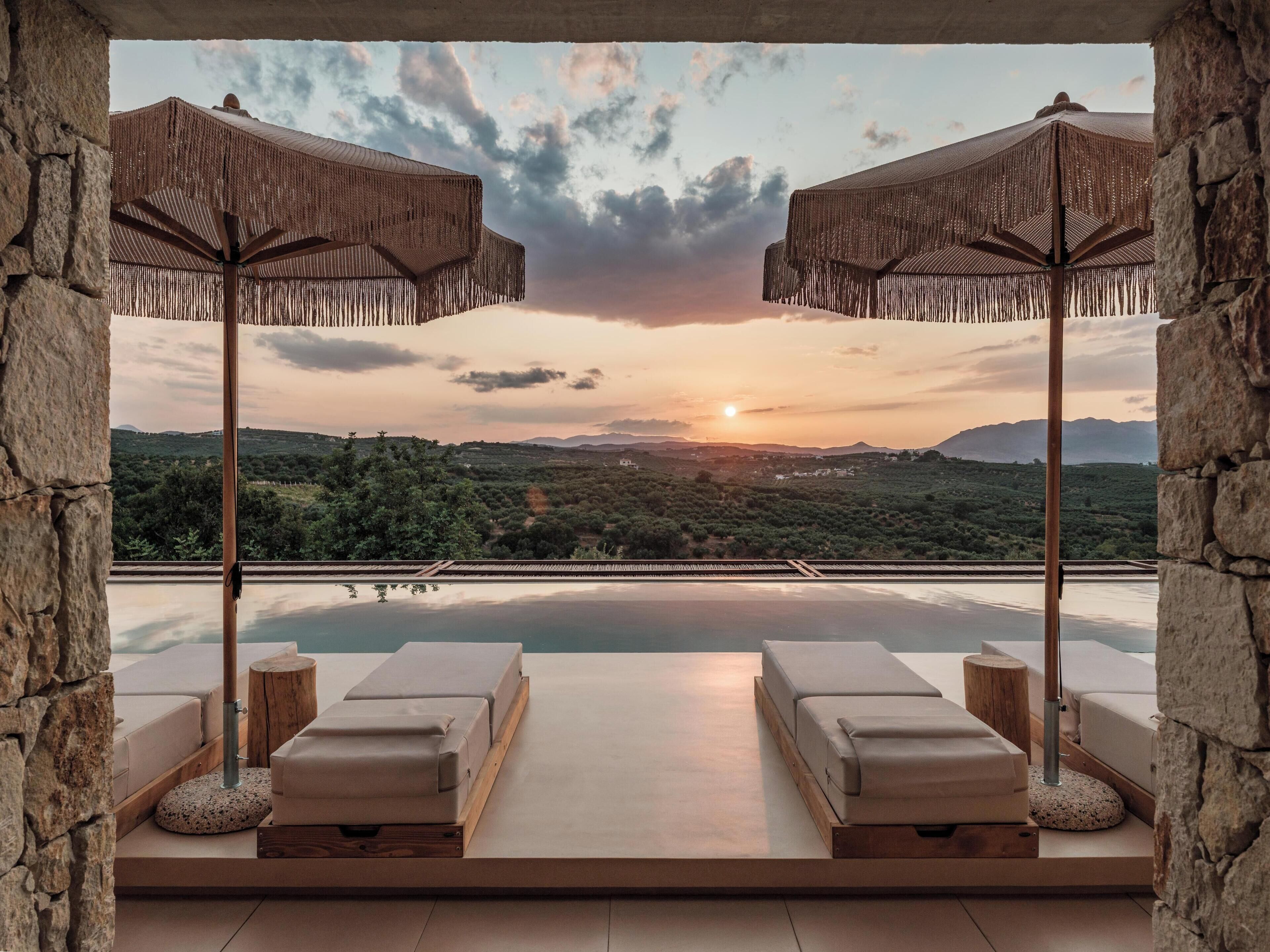 A terrace with two lounge chairs and two woven umbrellas, overlooking a pool and a scenic landscape during sunset.