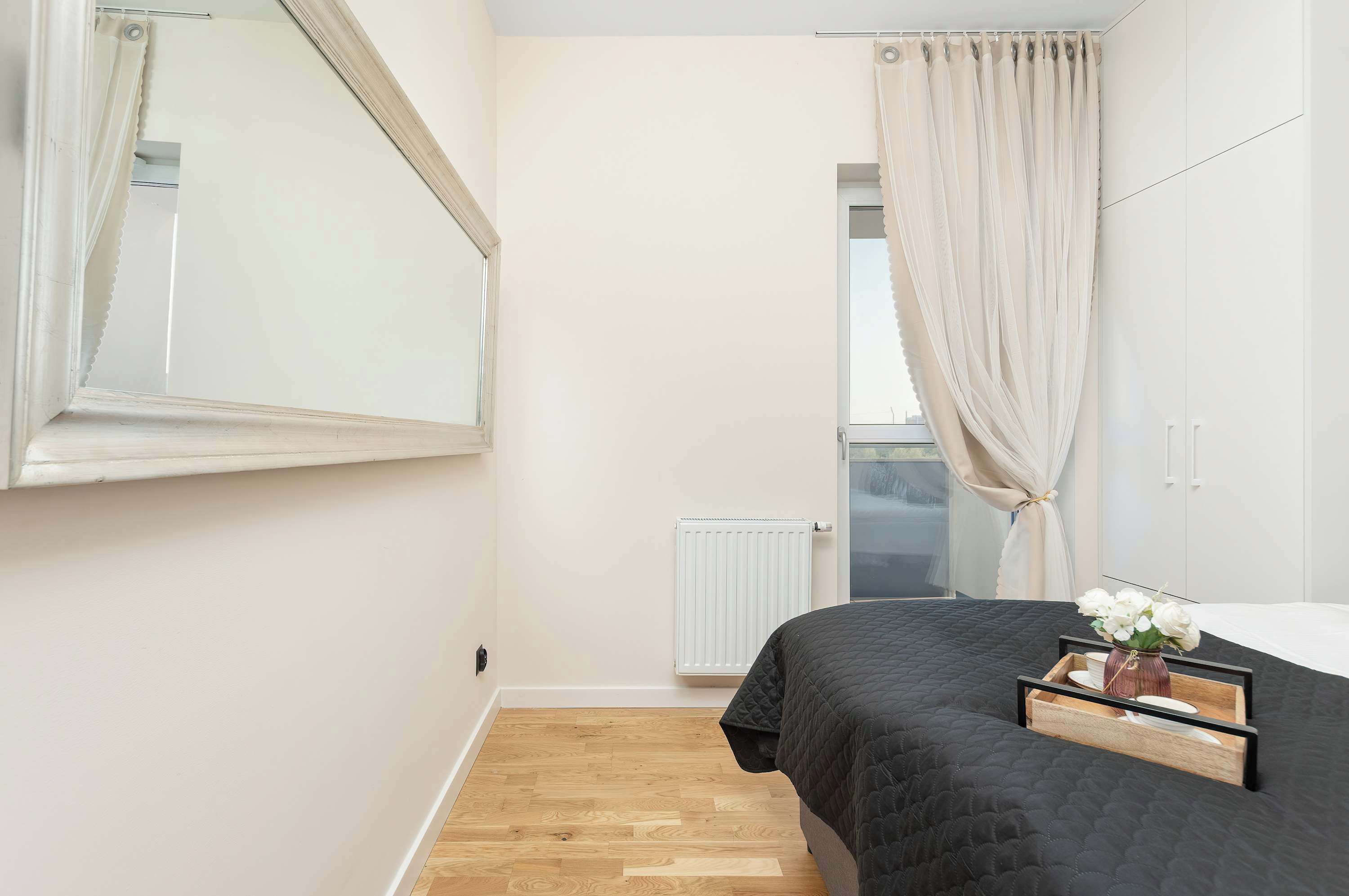 Een slaapkamer met een bed, een radiator, een raam met gordijnen en een spiegel.