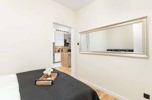 Um quarto com uma cama, uma mesa de cabeceira com um vaso de flores, um espelho grande e vista para a cozinha.