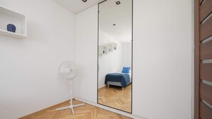 A bedroom with a large mirror, a bed with blue bedding, a white fan, and a wooden floor. - Practical Family Apartment | 2 Bedrooms | Close to the Airport (Warszawa)