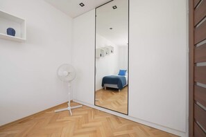 A bedroom with a large mirror, a bed with blue bedding, a white fan, and a wooden floor. - Practical Family Apartment | 2 Bedrooms | Close to the Airport (Warszawa)