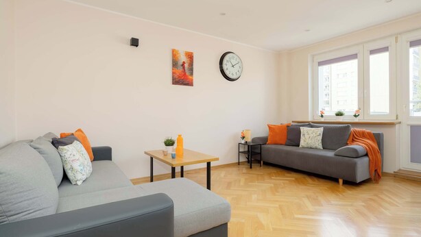 A modern living room with a grey sofa, a smaller grey couch, a wooden coffee table, and a large window with a view of buildings. - Practical Family Apartment | 2 Bedrooms | Close to the Airport (Warszawa)