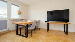 A dining area with a table set for four, a flat-screen TV mounted on a stand, and a window with a view of buildings. - Practical Family Apartment | 2 Bedrooms | Close to the Airport (Warszawa)