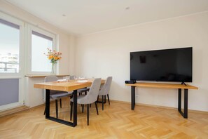 A dining area with a table set for four, a flat-screen TV mounted on a stand, and a window with a view of buildings. - Practical Family Apartment | 2 Bedrooms | Close to the Airport (Warszawa)