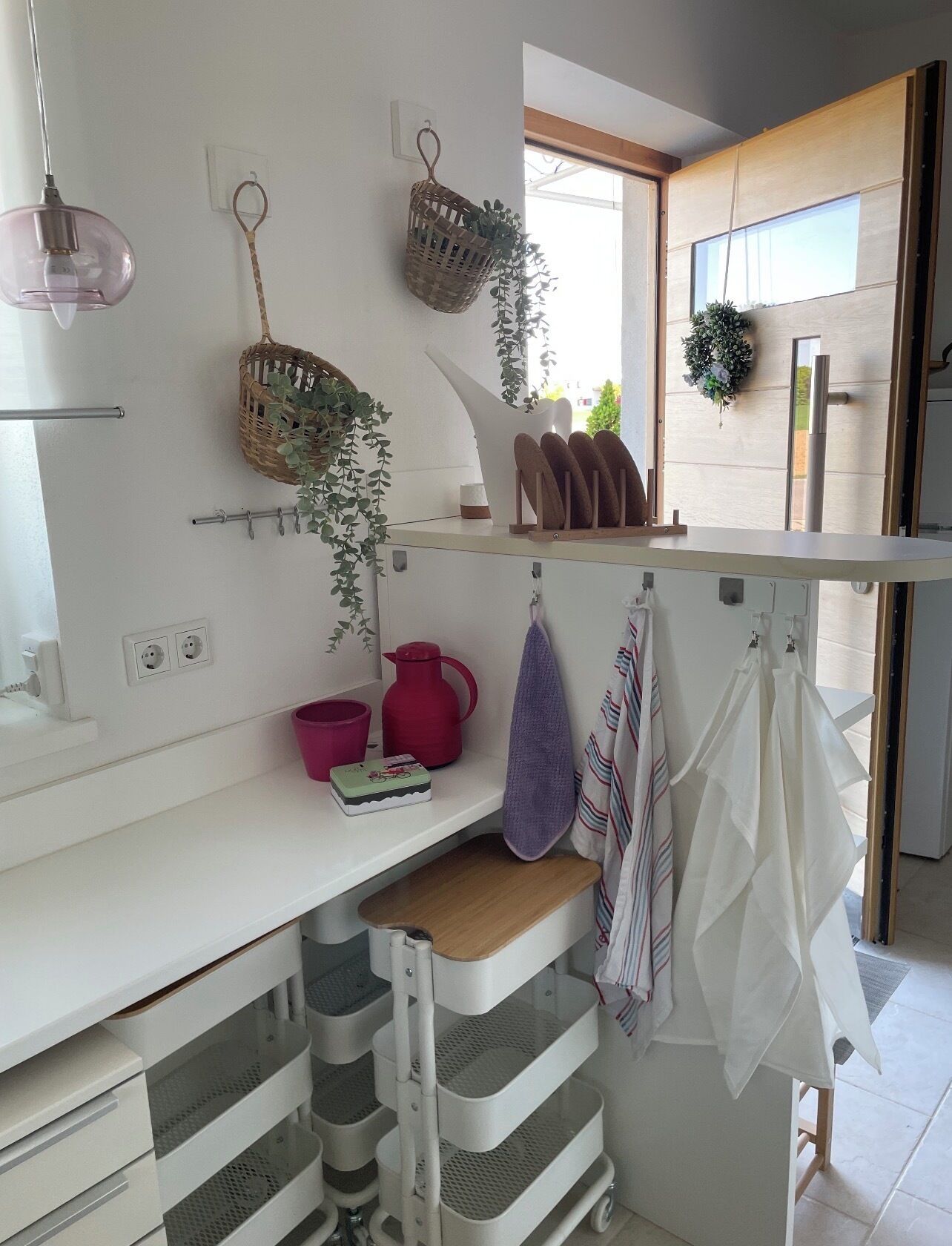 A modern kitchen with white cabinetry, hanging baskets with plants, a wooden countertop, and a window providing natural light.