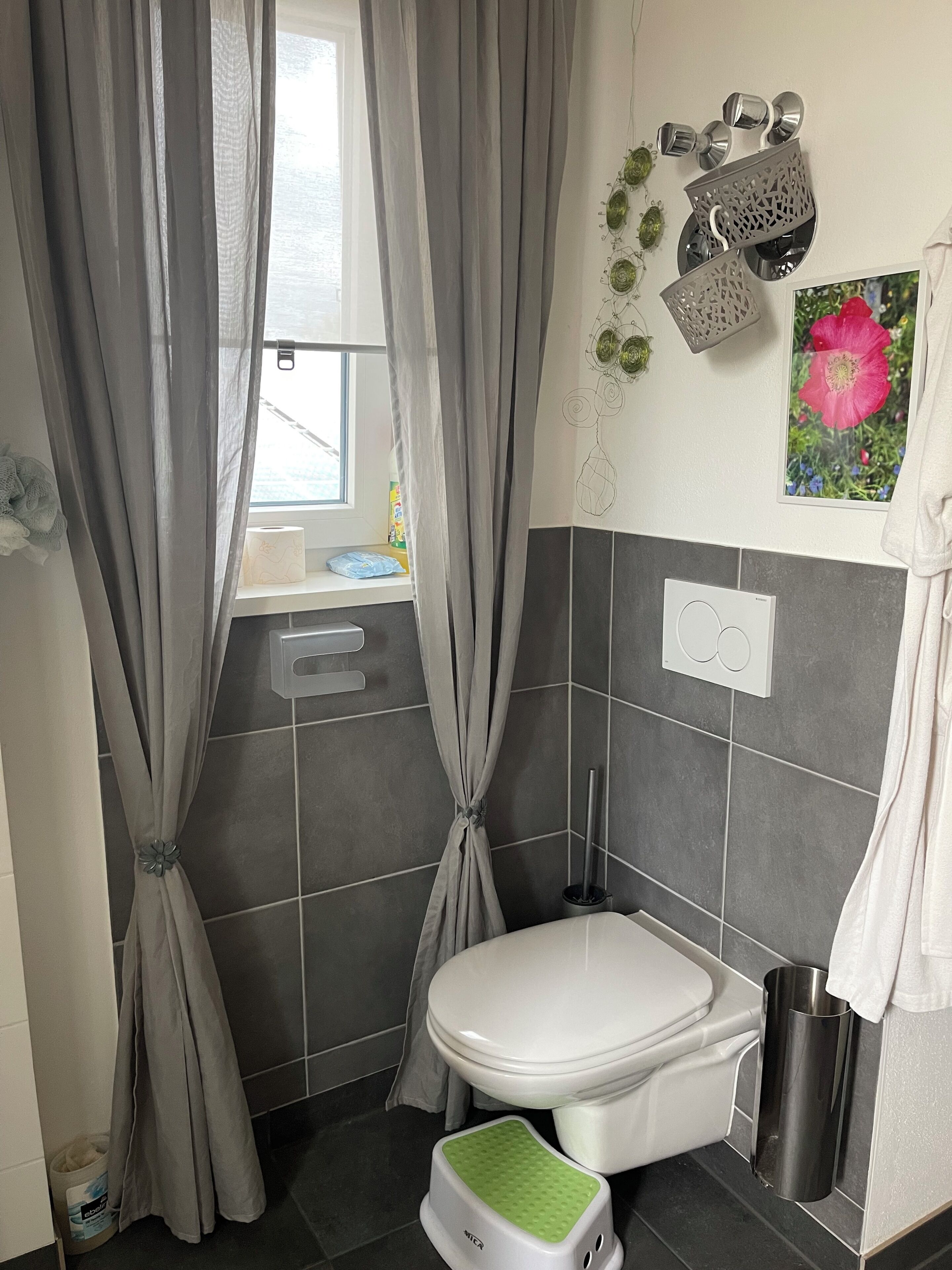 Un baño con inodoro, un taburete, un cubo de basura, una ventana con cortinas y una zona de ducha.