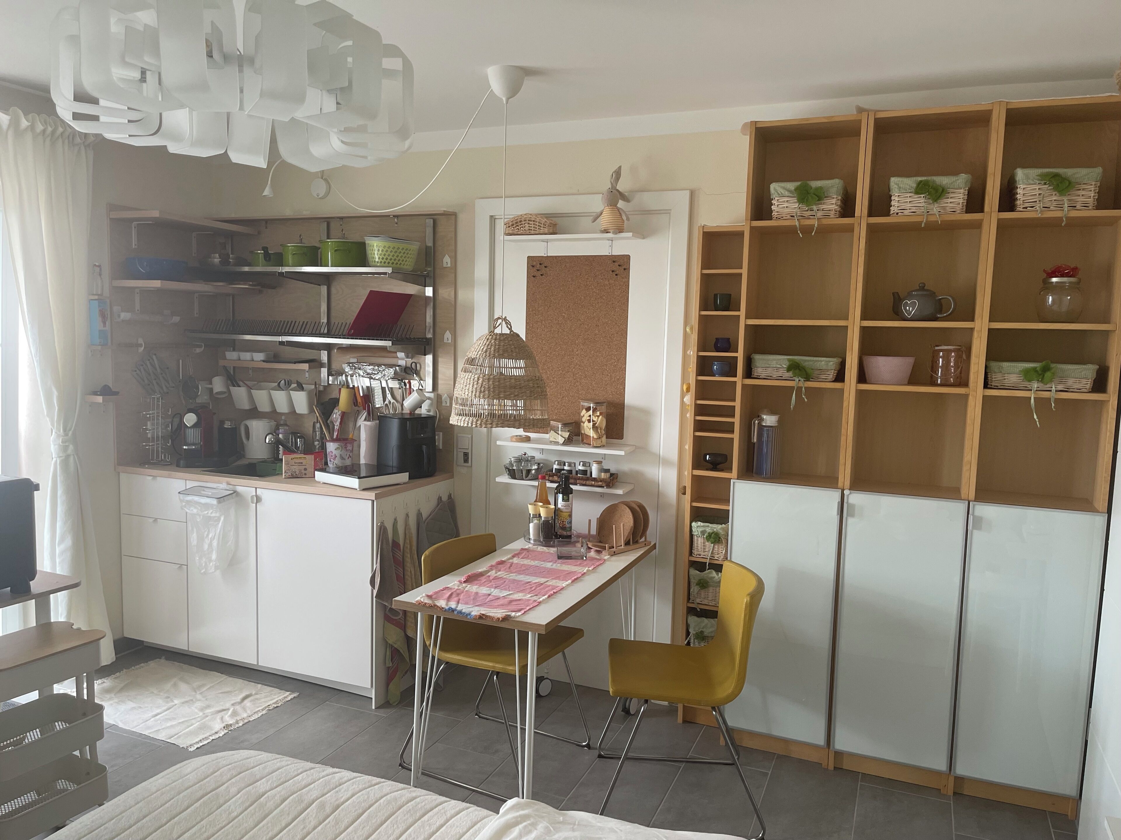A compact kitchen with a dining area, featuring a table with chairs, a hanging light fixture, and a wooden shelving unit with kitchenware.