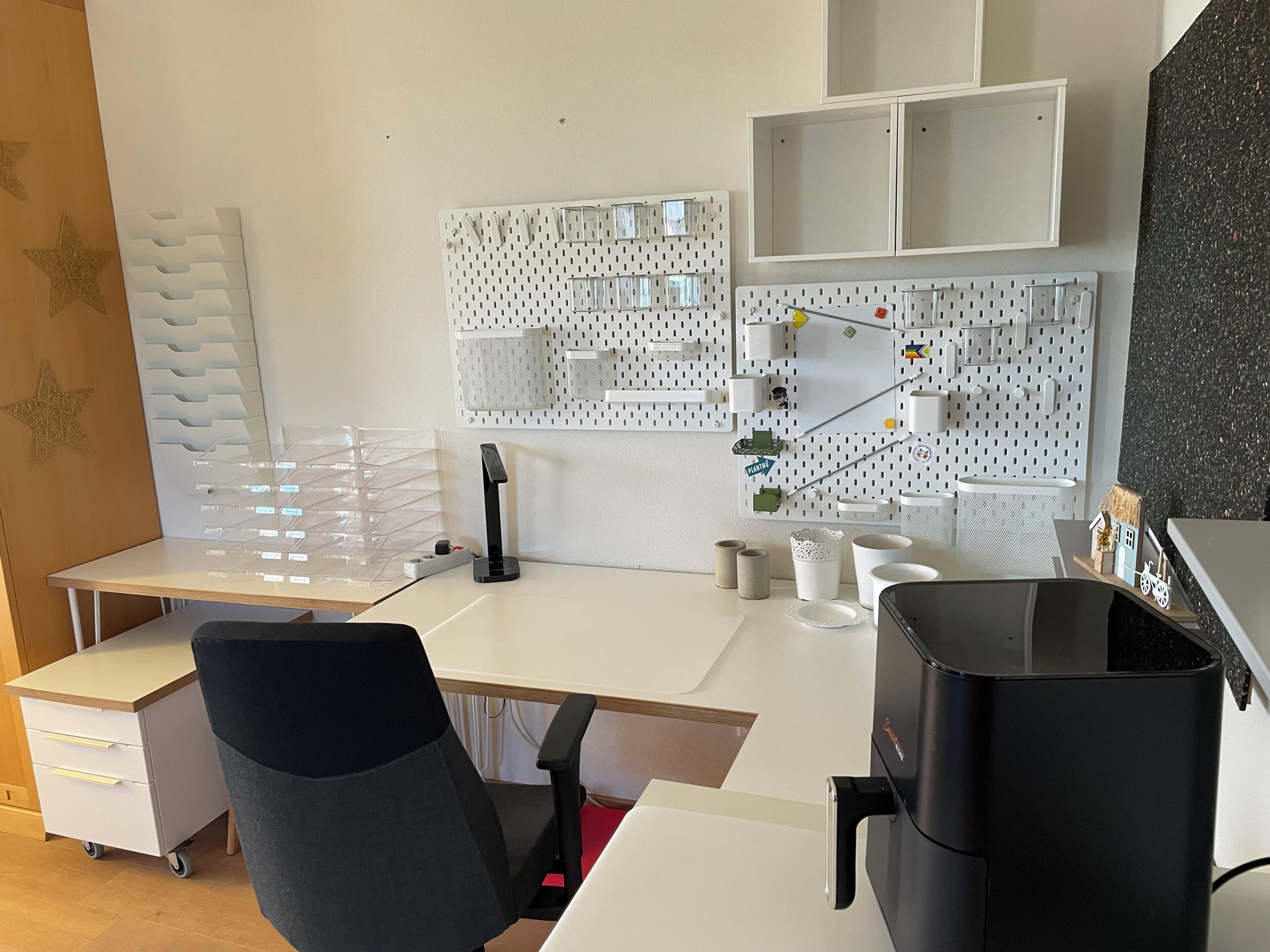Un espacio de oficina moderno con un escritorio blanco, una silla de oficina negra, una máquina de café y una pared de tableros con diversas soluciones de almacenamiento.