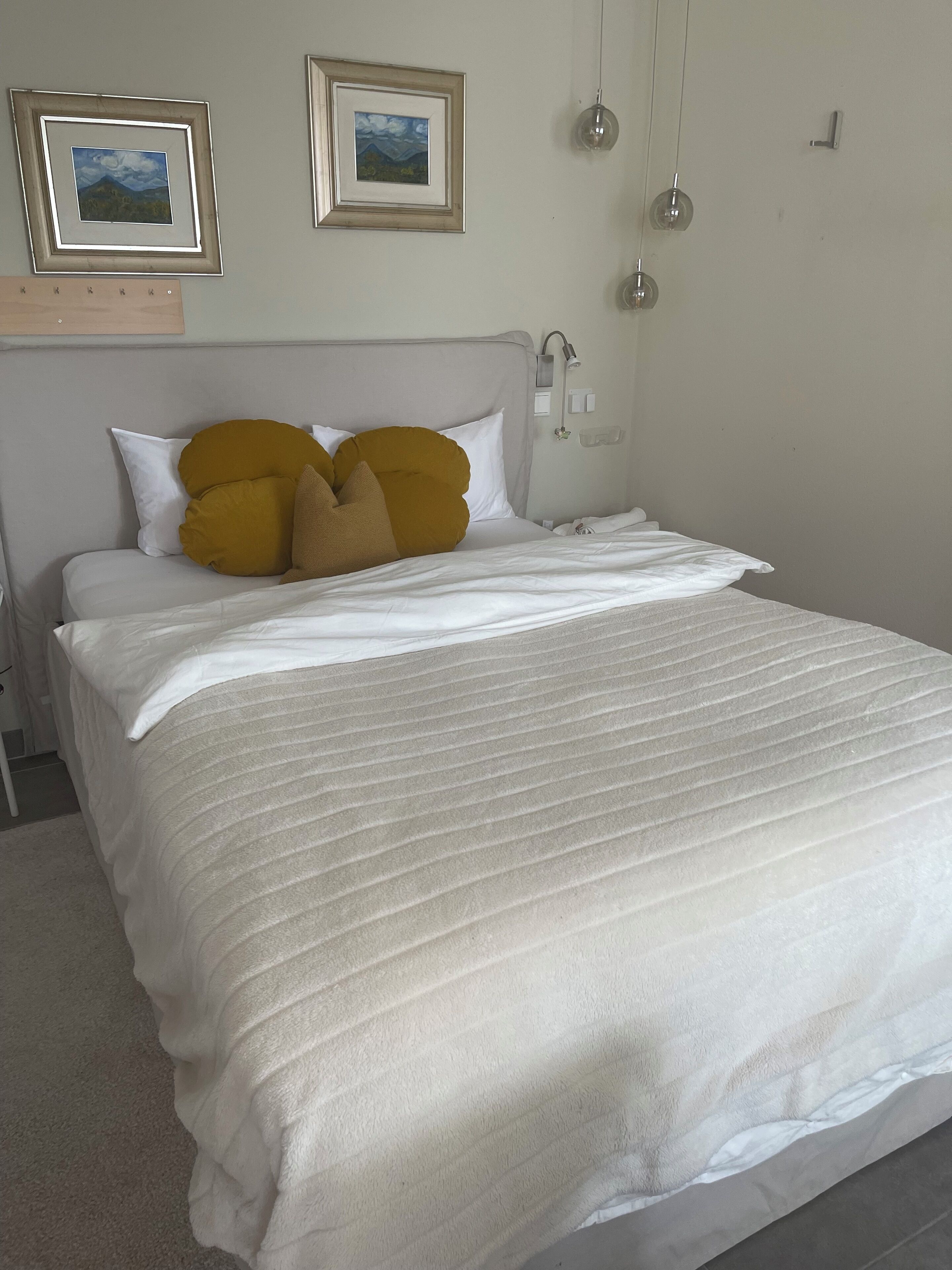 A neatly made bed with white bedding and mustard-colored pillows, a bedside table, and two framed pictures on the wall.