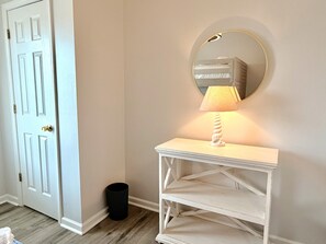 A hallway with a white door, a round mirror, a white console table with a lamp, and a black trash can. - Turtle Cottage: 3BR 2BA, Oceanfront, Rooftop Deck, Gazebo, Sleeps 7 (Oak Island)