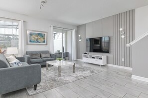 A modern living room with a grey sofa, a coffee table, a flat-screen TV, and a staircase leading to an upper level. - Spacious Family Retreat w/ Pvt Pool + BBQ at Champions Gate (Davenport)