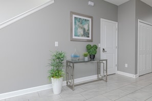 Un couloir avec une table à plateau de verre, des plantes en pot, une œuvre d’art encadrée et une porte blanche.