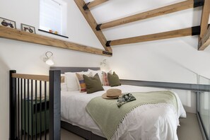 A bedroom with a sloped ceiling, a bed with multiple pillows, a bedside table, and a window with blinds. - The Old Abattoir -  an apartment that sleeps 2 guests  in 1 bedroom (Chipping Campden)