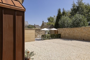 A courtyard with a gravel surface, stone walls, and a wooden building. There are two umbrellas providing shade. - The Old Abattoir -  an apartment that sleeps 2 guests  in 1 bedroom (Chipping Campden)