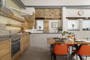 A modern kitchen with a stone wall, wooden cabinets, and a dining area with orange chairs. - The Old Abattoir -  an apartment that sleeps 2 guests  in 1 bedroom (Chipping Campden)
