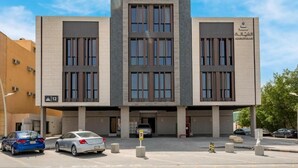 A modern building with a central entrance, multiple windows, and a parking area with cars.