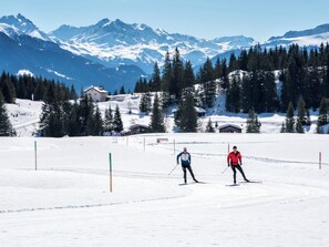 A snowy ski slope with two skiers, a mountain backdrop, and a small building in the distance.