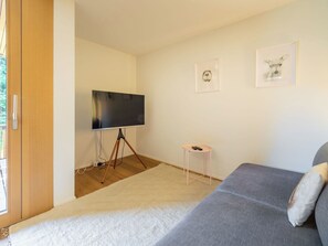 Ein modernes Wohnzimmer mit einem grauen Sofa, einem Flachbildfernseher auf einem Stativ, einem kleinen weißen Beistelltisch und zwei gerahmten Bildern an der Wand.