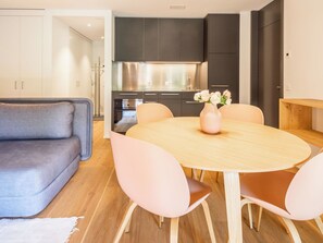 A modern kitchen with dark cabinets and stainless steel appliances, a round dining table with chairs, and a grey sofa in a living area.